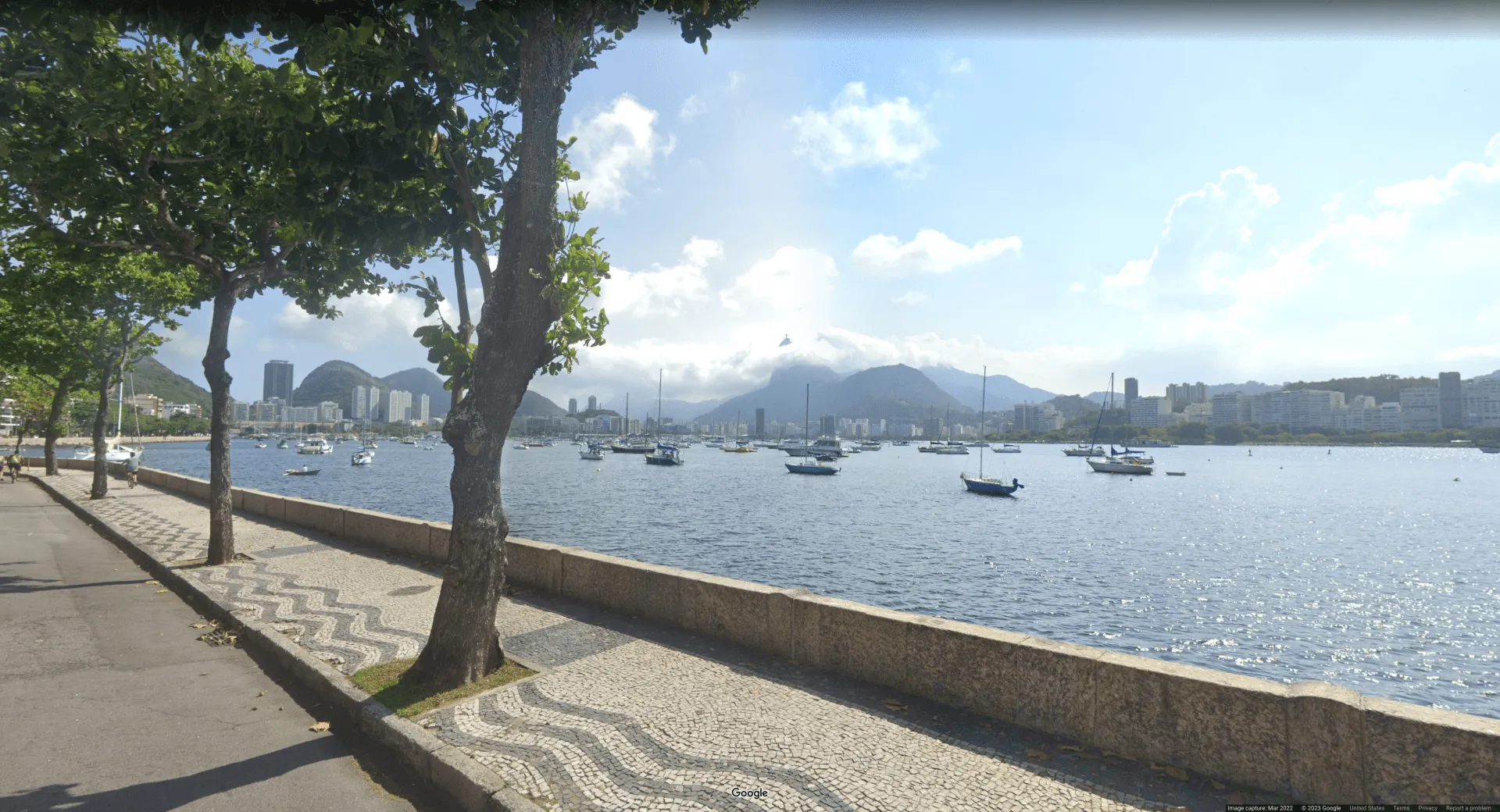 Picture of a waterfront walkway near a sea, with boats in a local harbor with overcast skies. A small statue is visible in the background, peeking above the clouds